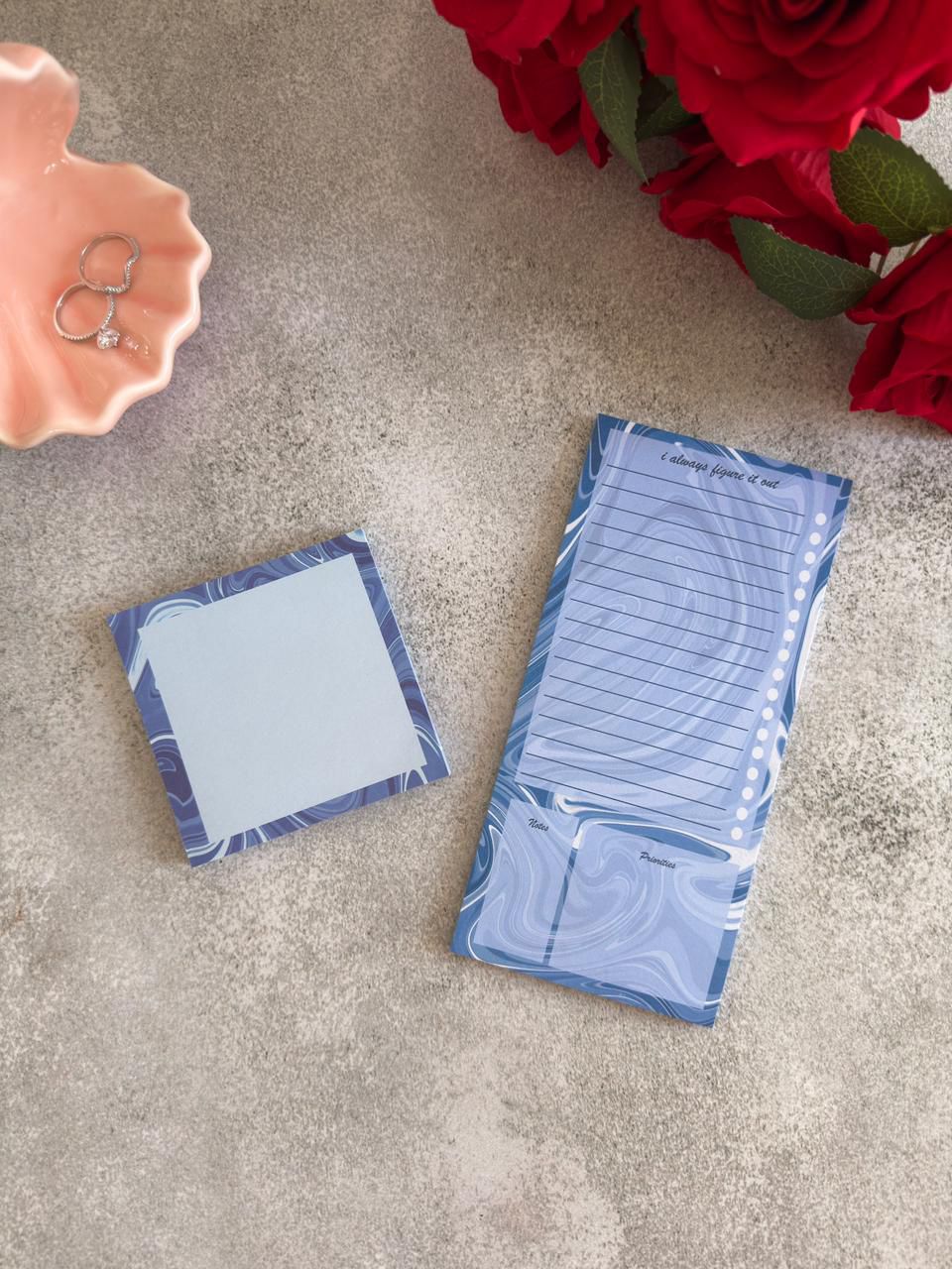 Two blue notepads on a textured surface with red flowers and a pink dish in the background.